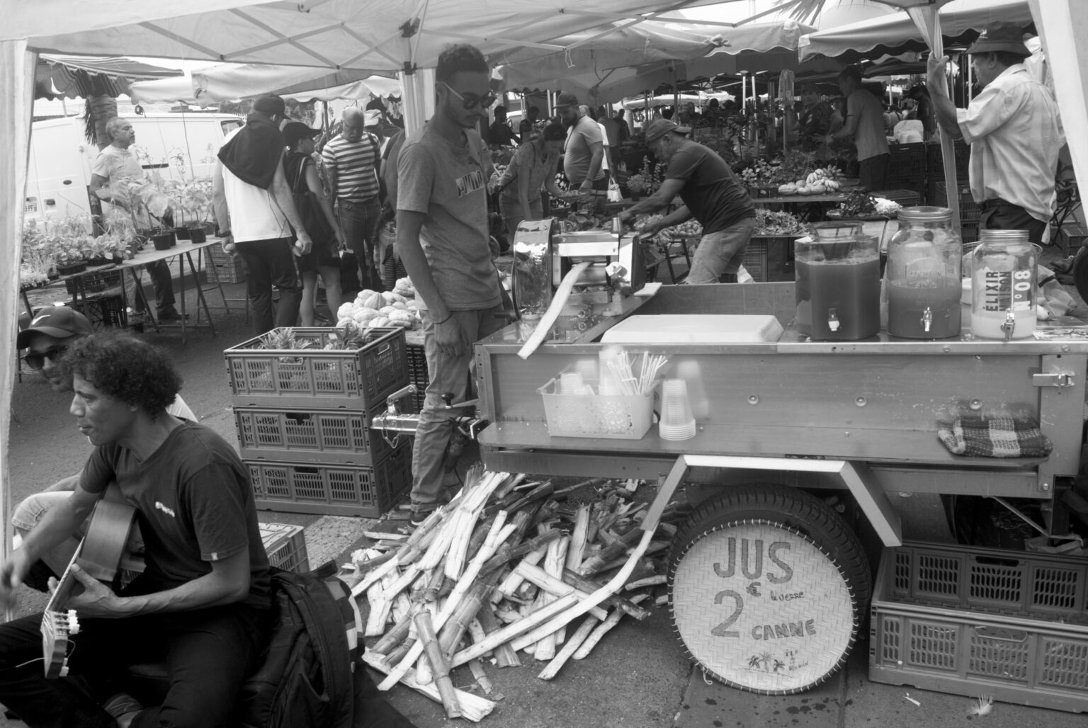市場のサトウキビ搾り機と「JUS 2€ le litre CANNE PÉI」の札、そばでギターを弾く人（白黒写真）