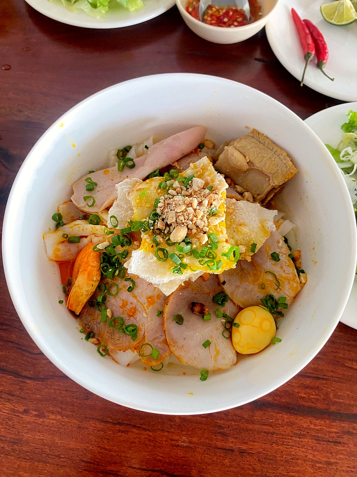 白い丼に太麺、練りもの、海老、うずらの卵、砕いたピーナッツ