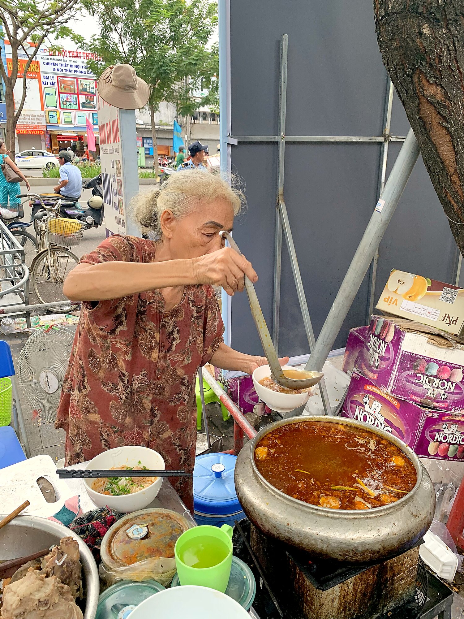 歩道の屋台で、年配の女性が大きな寸胴のスープをかき混ぜている。フエのドンバ市場
