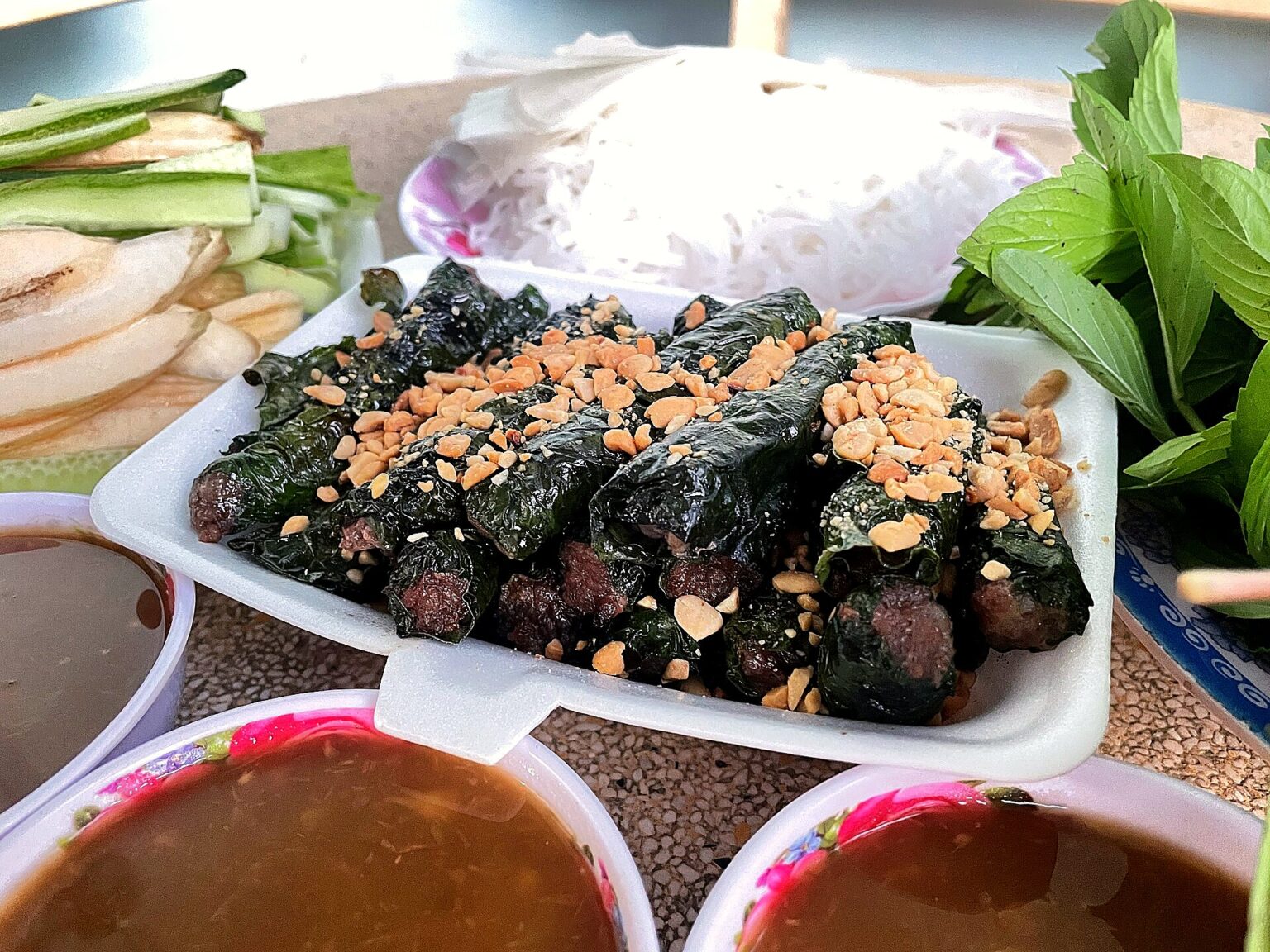 白い角皿に葉巻きの肉、砕いたピーナッツ。奥に細い米麺と香草、手前にたれの椀が二つ