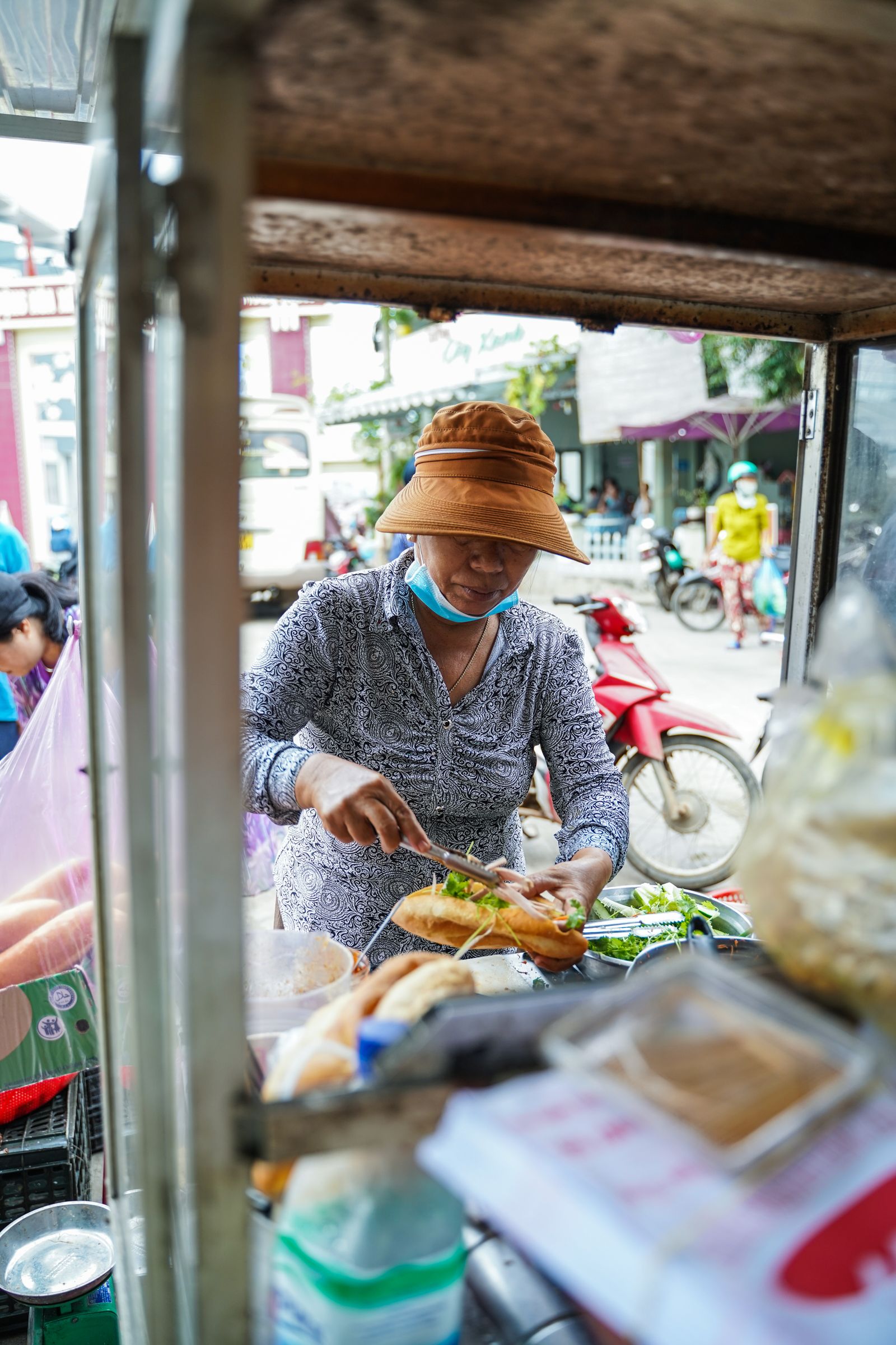 帽子をかぶった売り手が、バゲットを並べた屋台でバインミーに具を詰めている