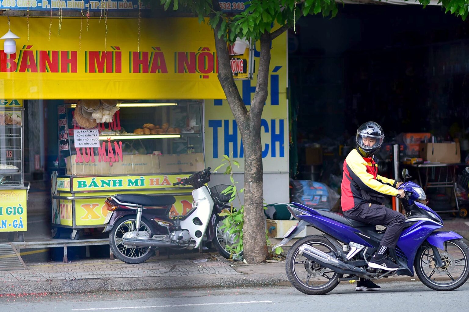 「BÁNH MÌ HÀ NỘI」の看板が並ぶ通りをバイクが走り抜ける（ベトナム国内・撮影地の詳細は不明）
