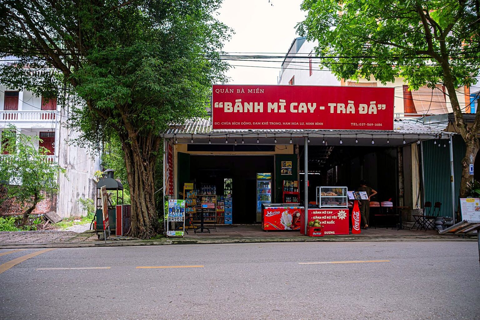木立の下、「BÁNH MÌ CAY - TRÀ ĐÁ」の赤い看板を掲げた店構え（ベトナム北部）