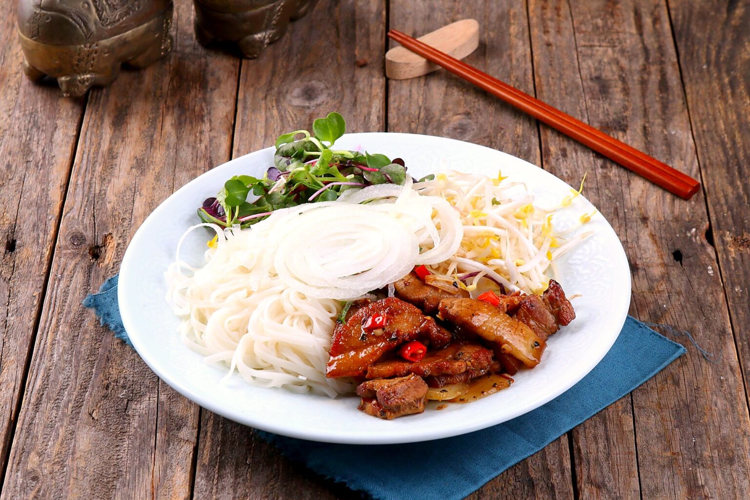 白い皿に、米麺、焼き豚肉、にんじんの甘酢漬け、香草が盛られている