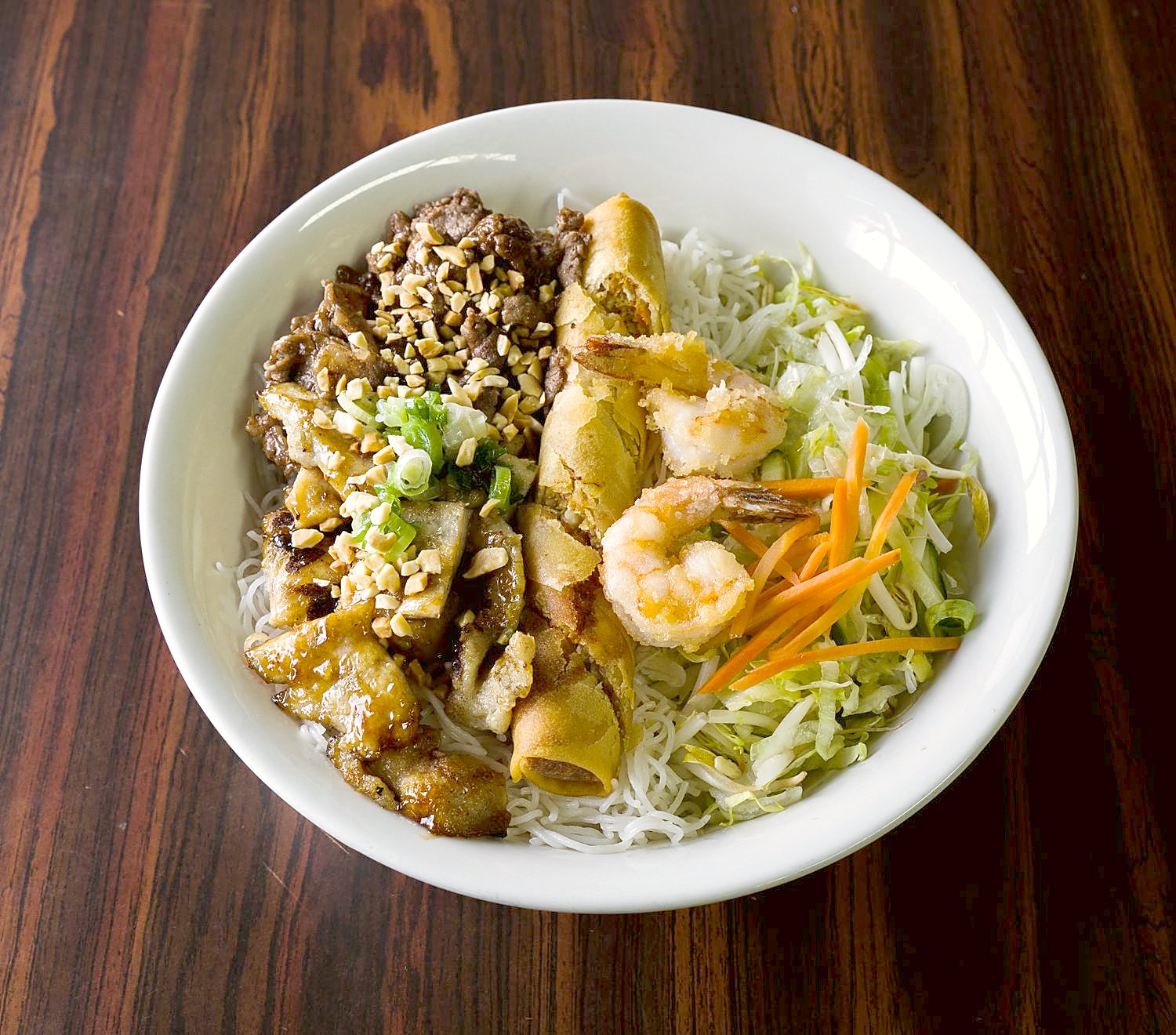 濃い色の木の卓に置いた白い丼。細い米麺の上に焼いた豚肉、海老、揚げ春巻き、なます