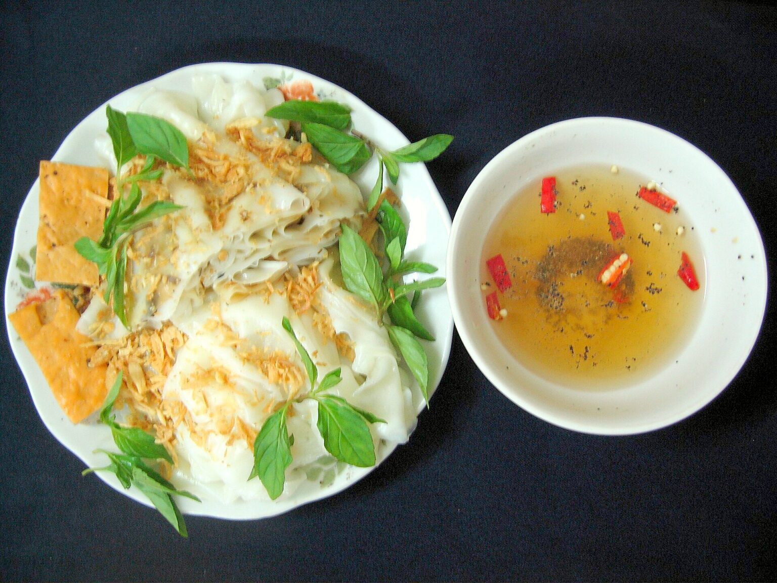 白い皿に、具を包まずに畳んだだけの薄皮と揚げもの、香草。となりに魚醤だれの椀。ハノイのタインチー風