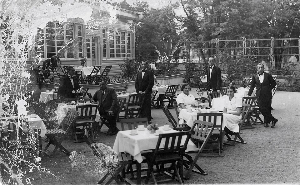 1930年代、白いクロスを掛けたテーブルと折りたたみ椅子が並ぶ屋外テラスと、給仕たちのモノクロ写真