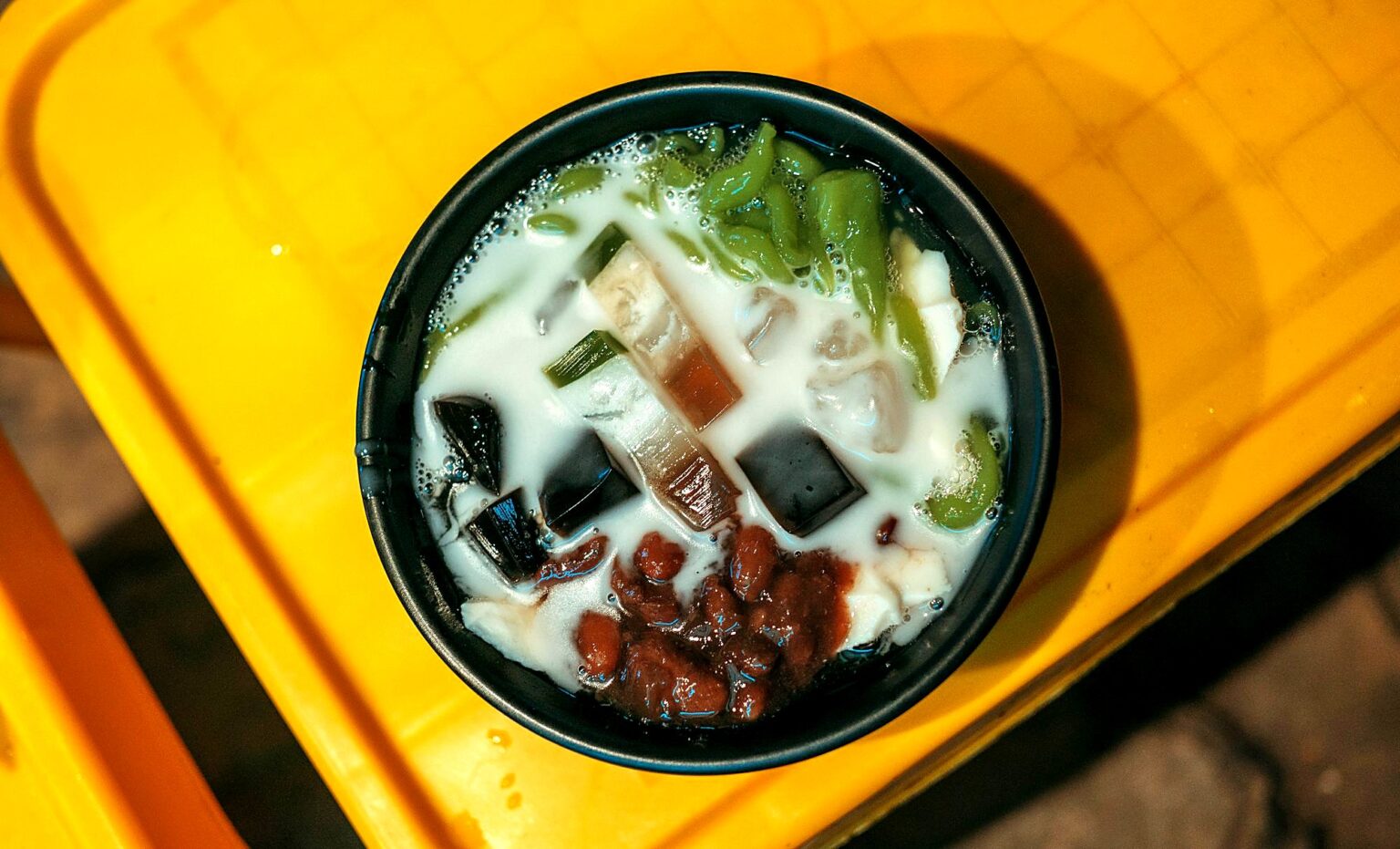 黄色い台の上の黒い器。ココナッツミルク、黒いゼリー、緑の麺、小豆、いもが入っている