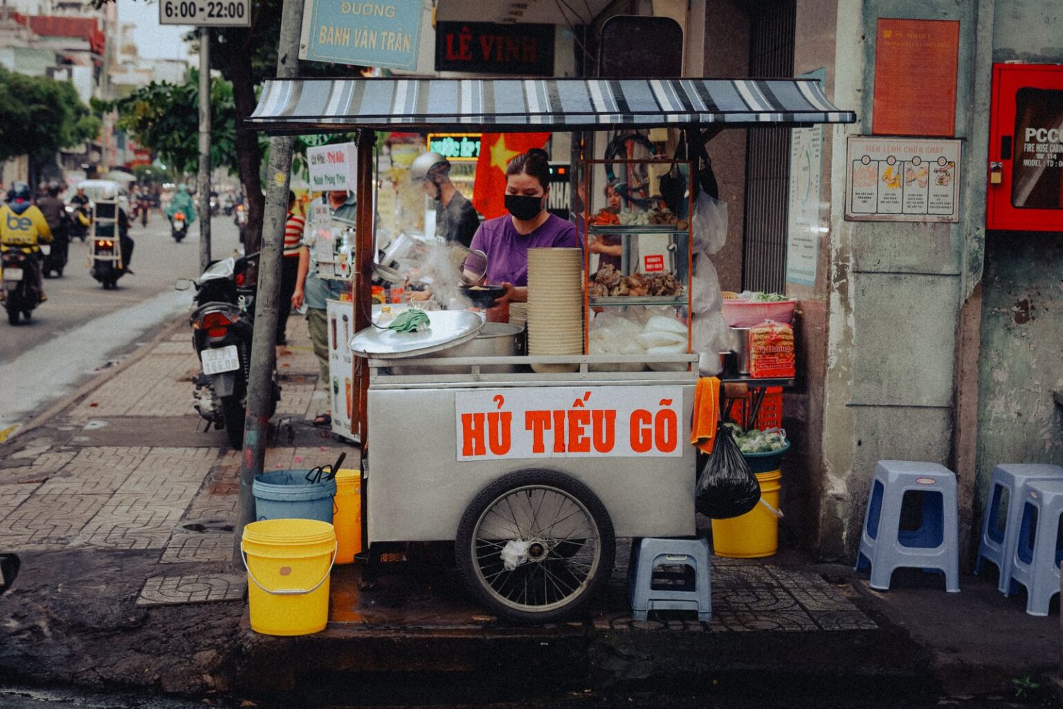 「HỦ TIẾU GÕ」と書かれた移動屋台のカート。歩道に青いプラ椅子、売り手