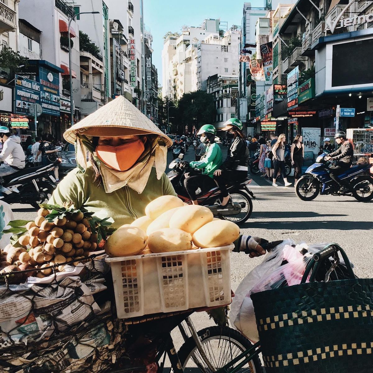 ノンラーの女性が自転車にパンと果物を積んで街路を行く。背景に高層ビル