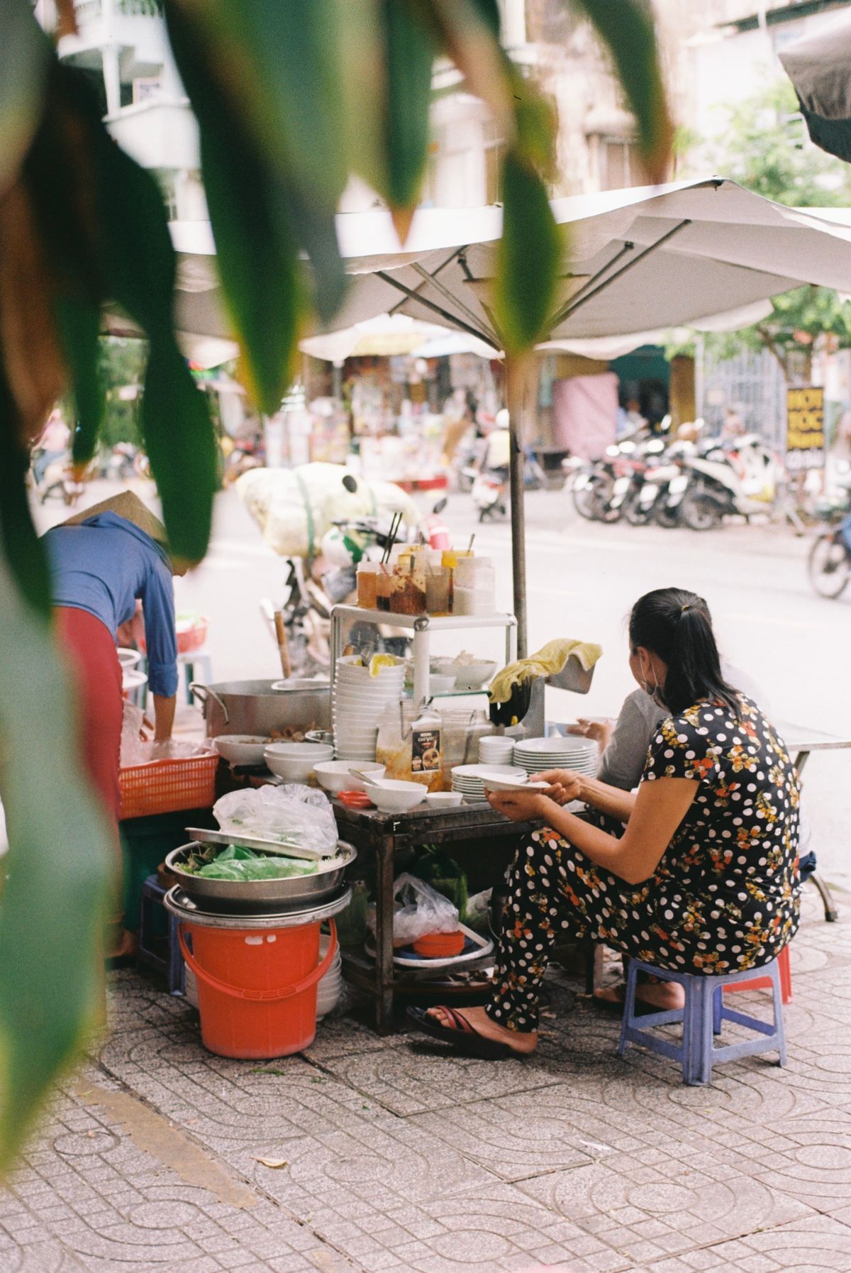 昼の歩道。パラソル下の屋台で座って食べる女性、前景に緑の葉