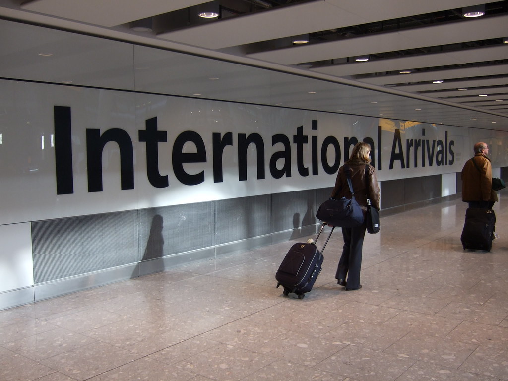 ヒースロー空港の「International Arrivals」の壁文字とスーツケースを引く旅行者