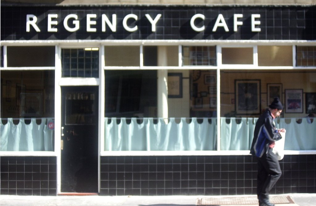 Regency Cafe の黒タイルの外観と白い「REGENCY CAFE」の文字、前を歩く人