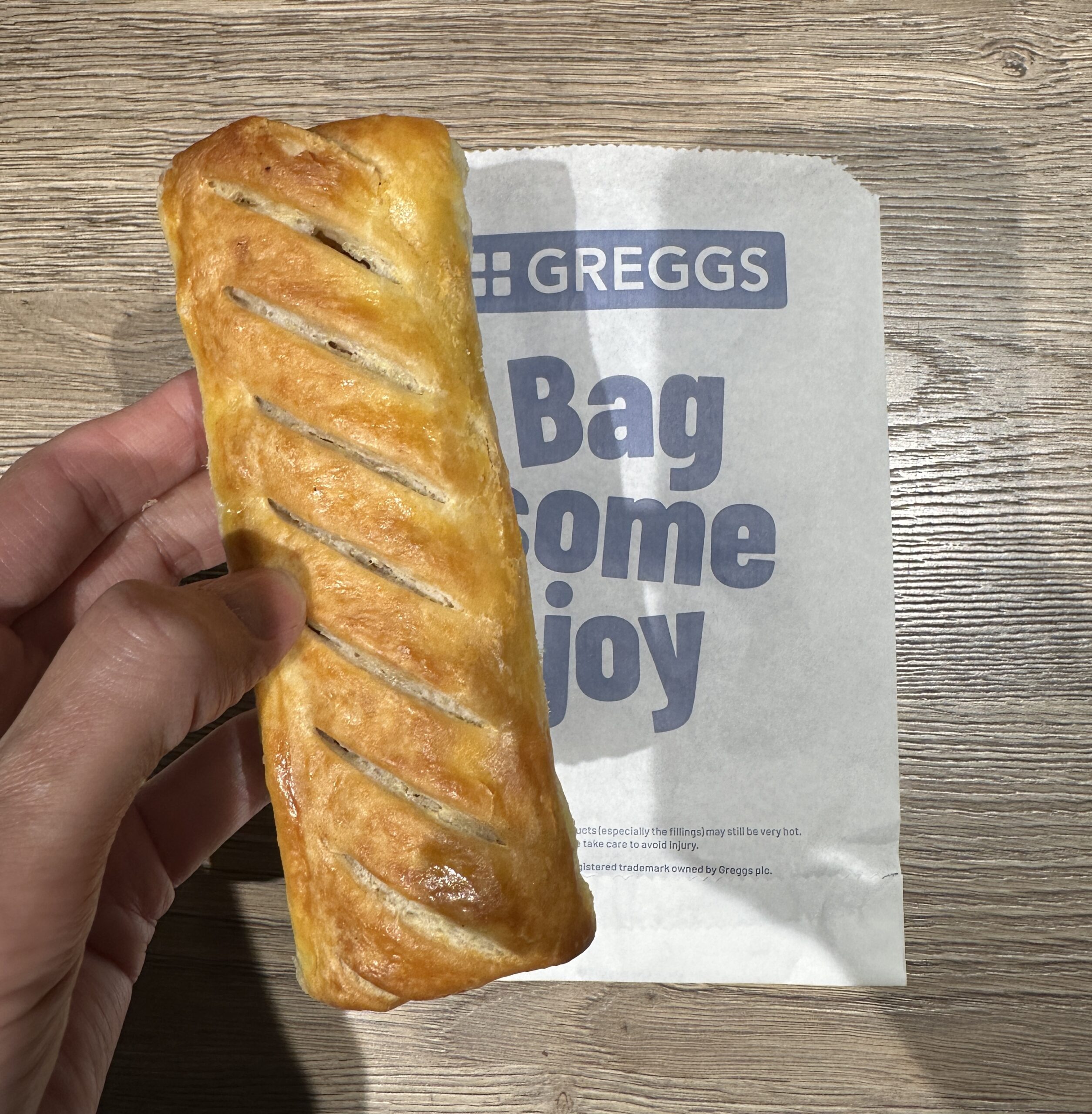 Greggs のソーセージ・ロールを手に持ち、Greggs の紙袋を背景にしたところ
