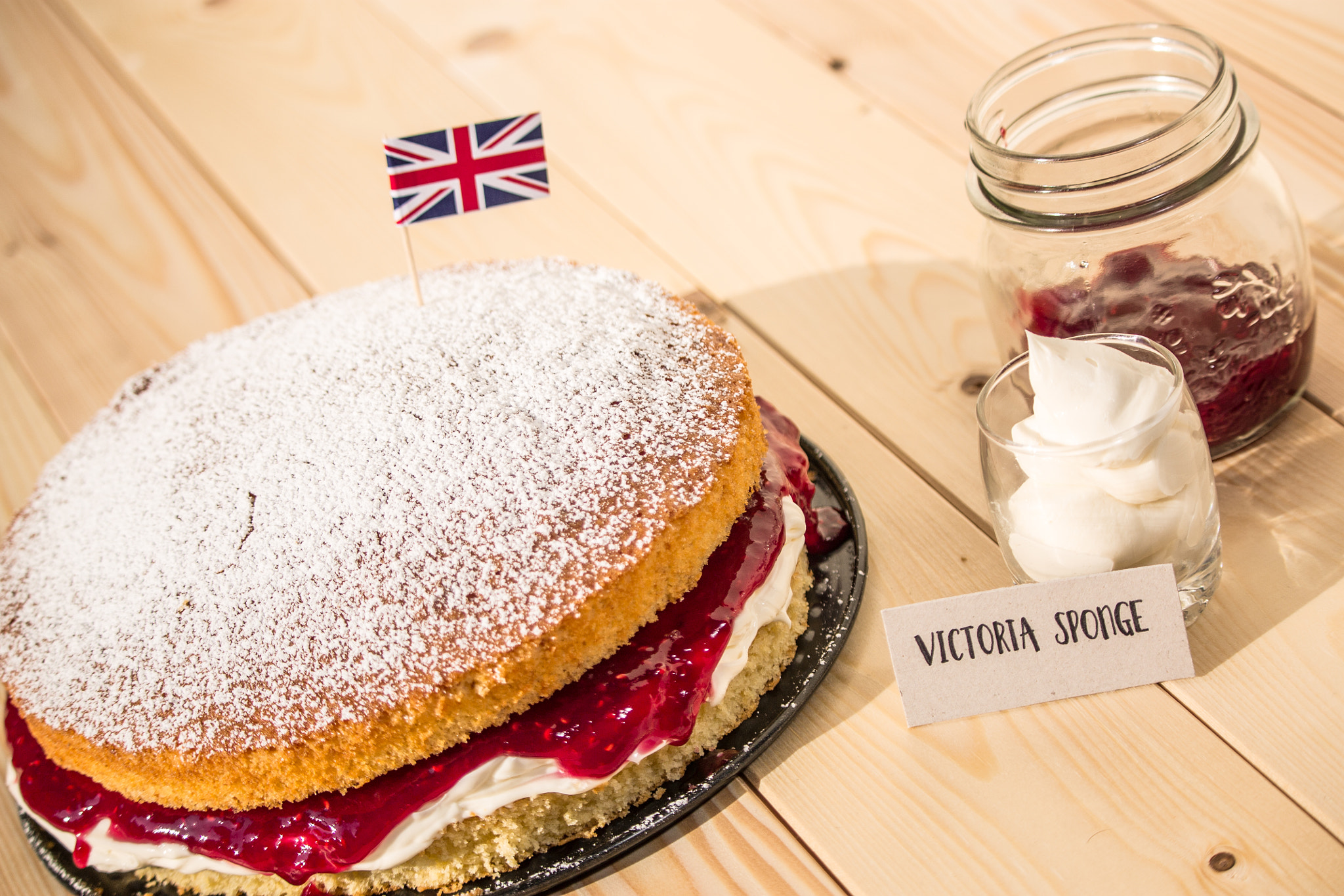 粉砂糖をふったヴィクトリア・スポンジのホールと英国旗の小旗、名札「Victoria sponge」