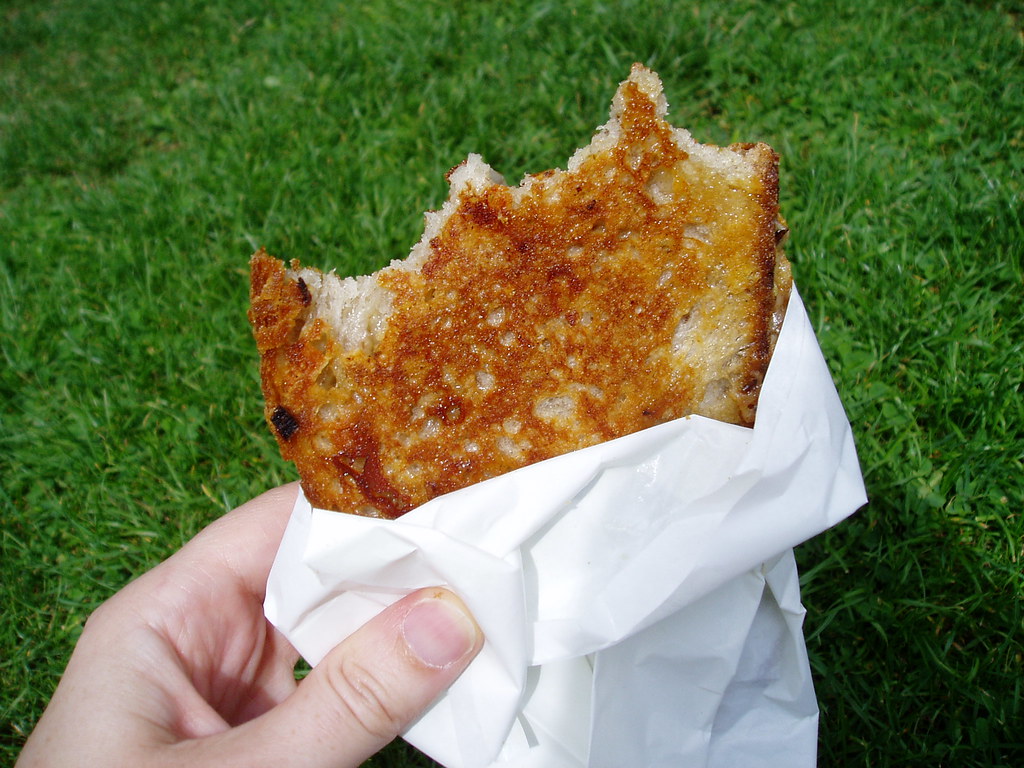 紙に包んだ焼き色の濃いチーズ・トースティーを手に持つ（芝生の上）