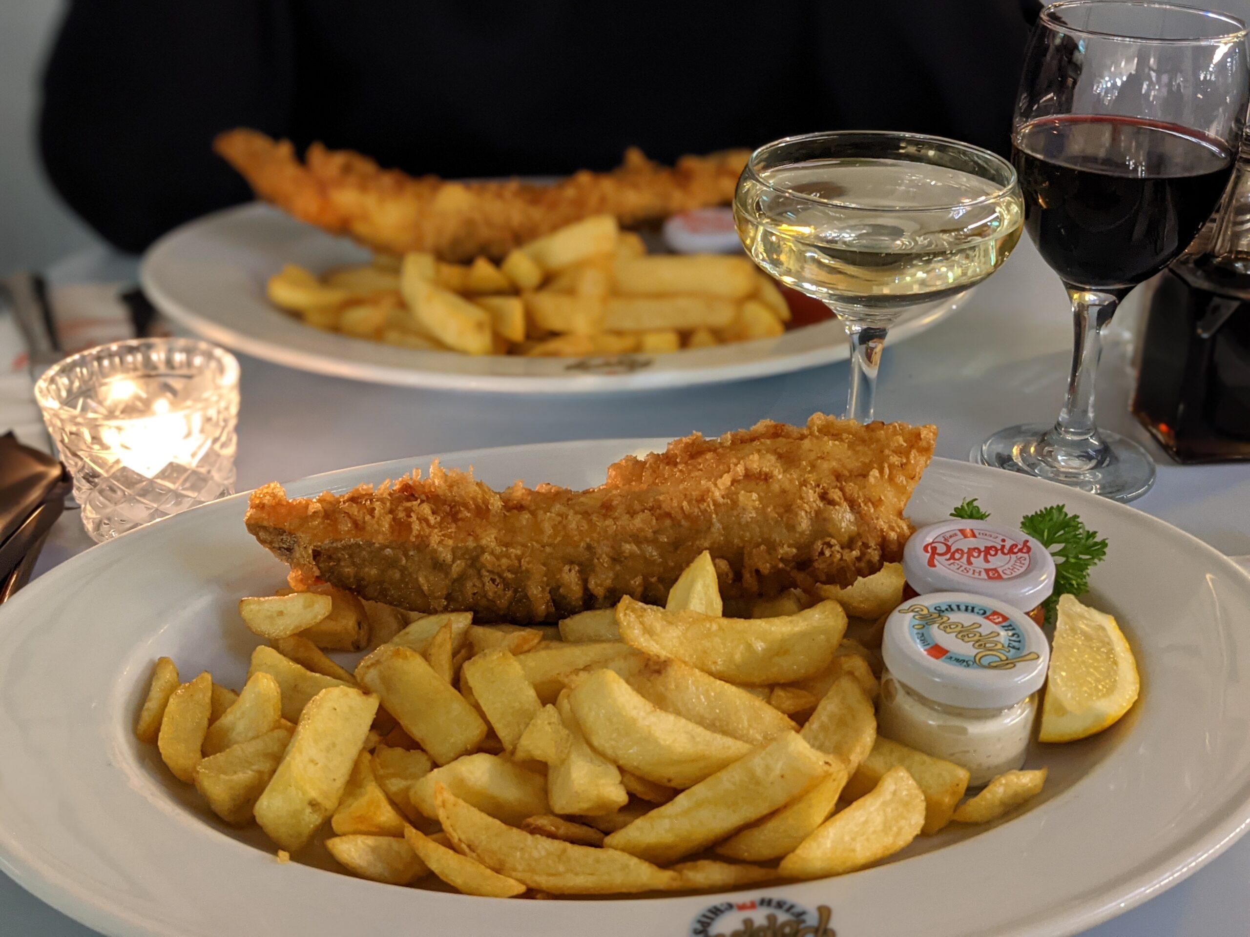 ポピーズ（スピタルフィールズ）のフィッシュ＆チップス。大きな衣の魚、太いチップス、レモンとタルタルソース、奥に白ワイン