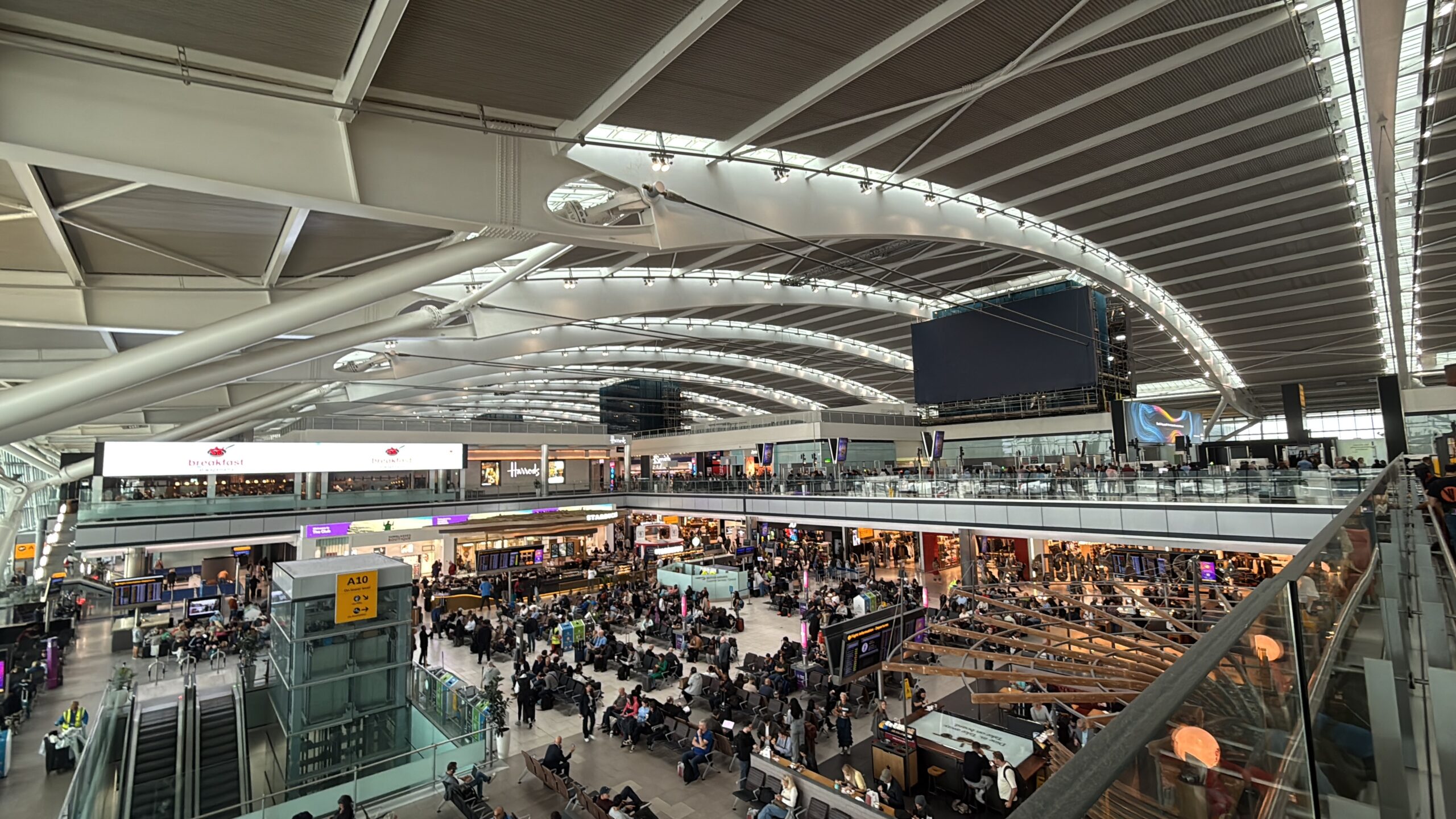 ヒースロー空港第5ターミナルの出発ロビー。波打つ白い大屋根の下に店とベンチ、大勢の旅客