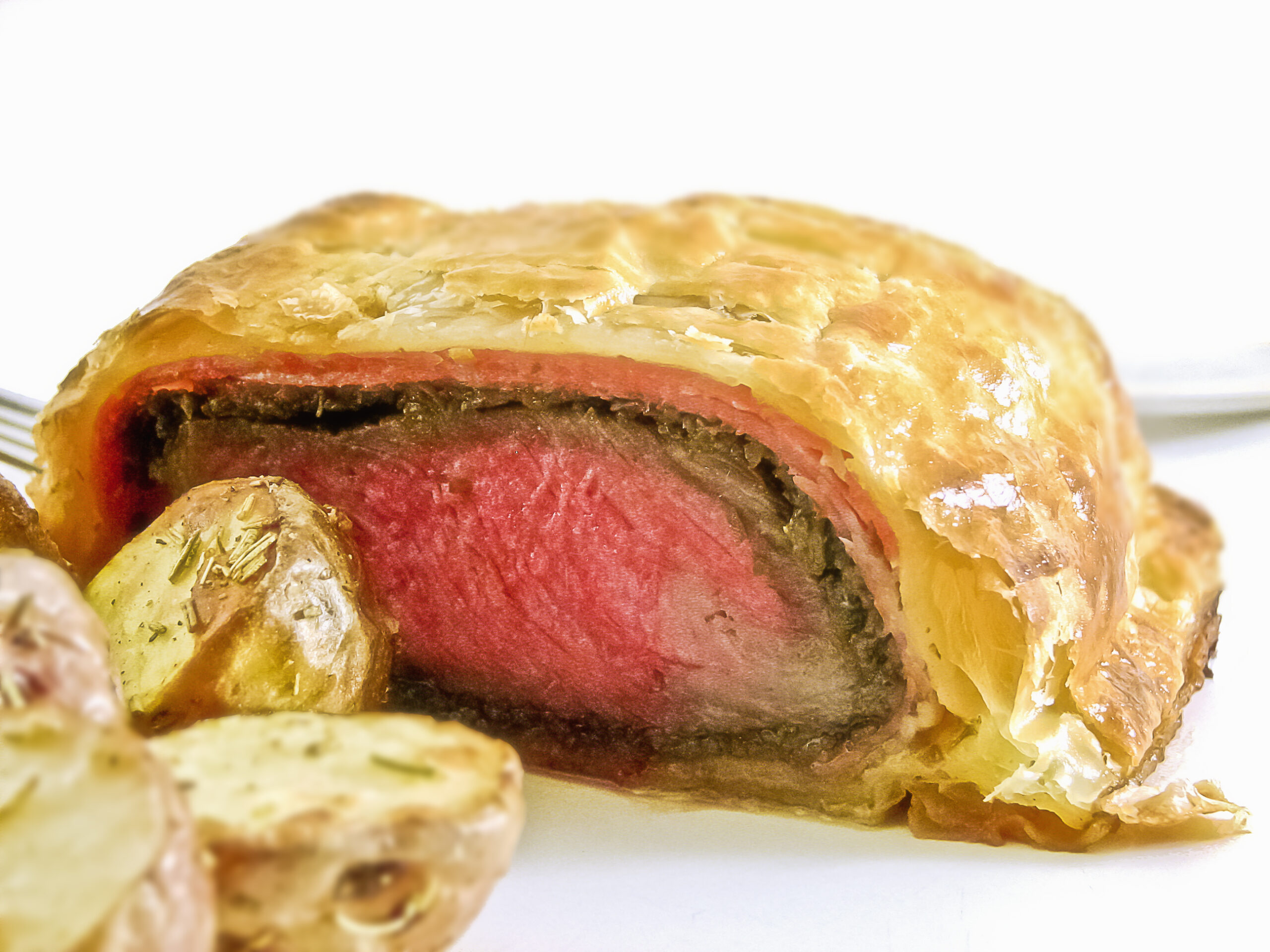 ビーフ・ウェリントンの断面の寄り。ロゼ色の牛肉、緑のデュクセル、パイ皮、手前にローストポテト