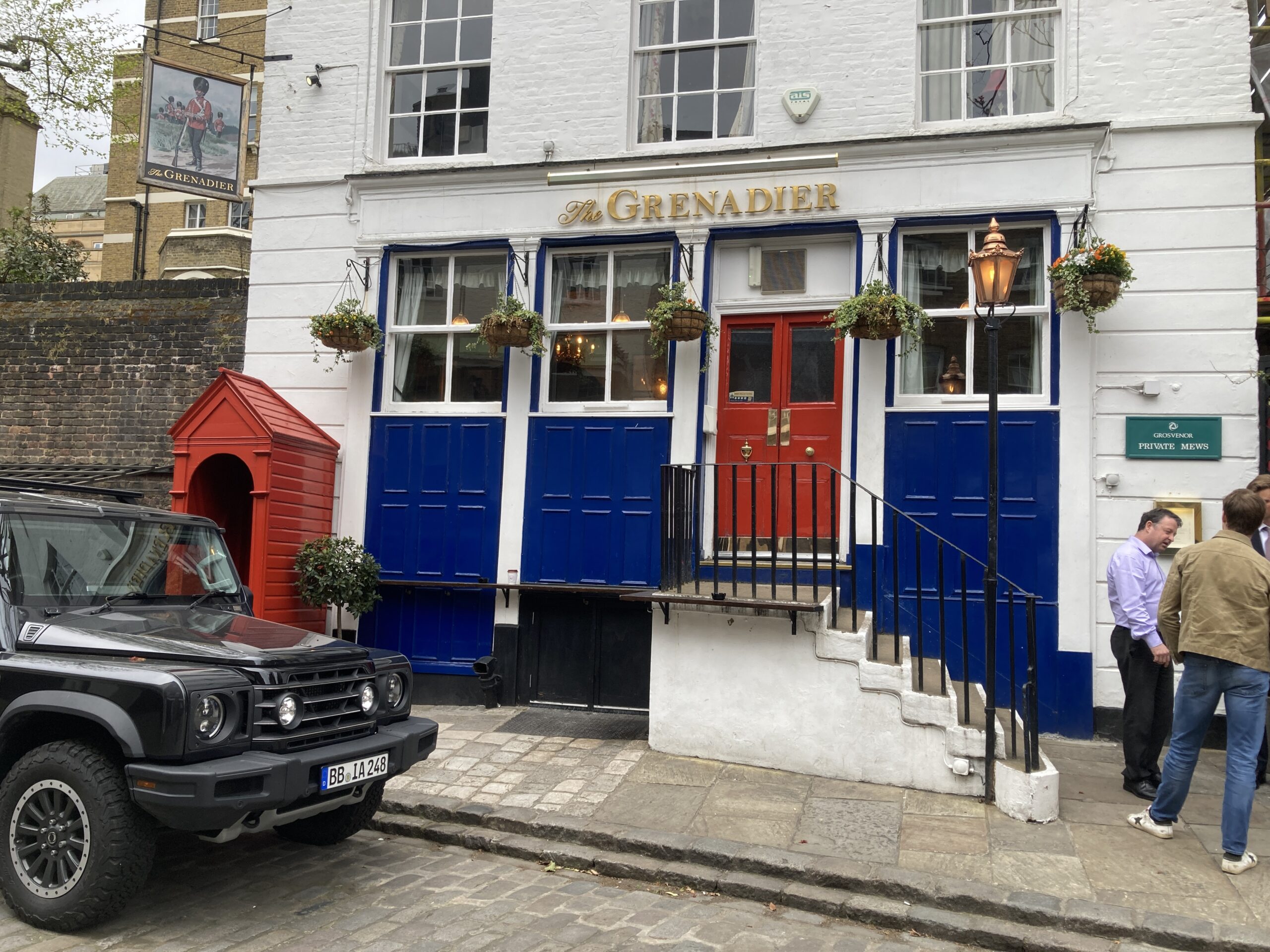 石畳の路地に建つ白いパブThe Grenadier。青い腰板、赤い扉、入口脇の赤い衛兵小屋、店先の客