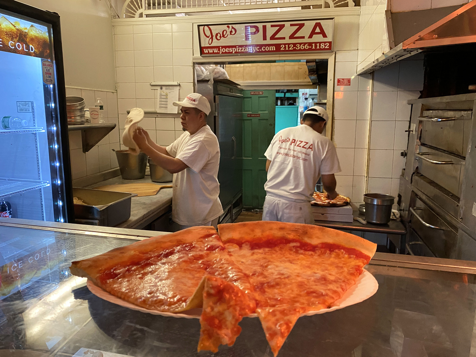 Joe's Pizza のカウンターに置かれたチーズ・ピザと奥で生地をのばす店員