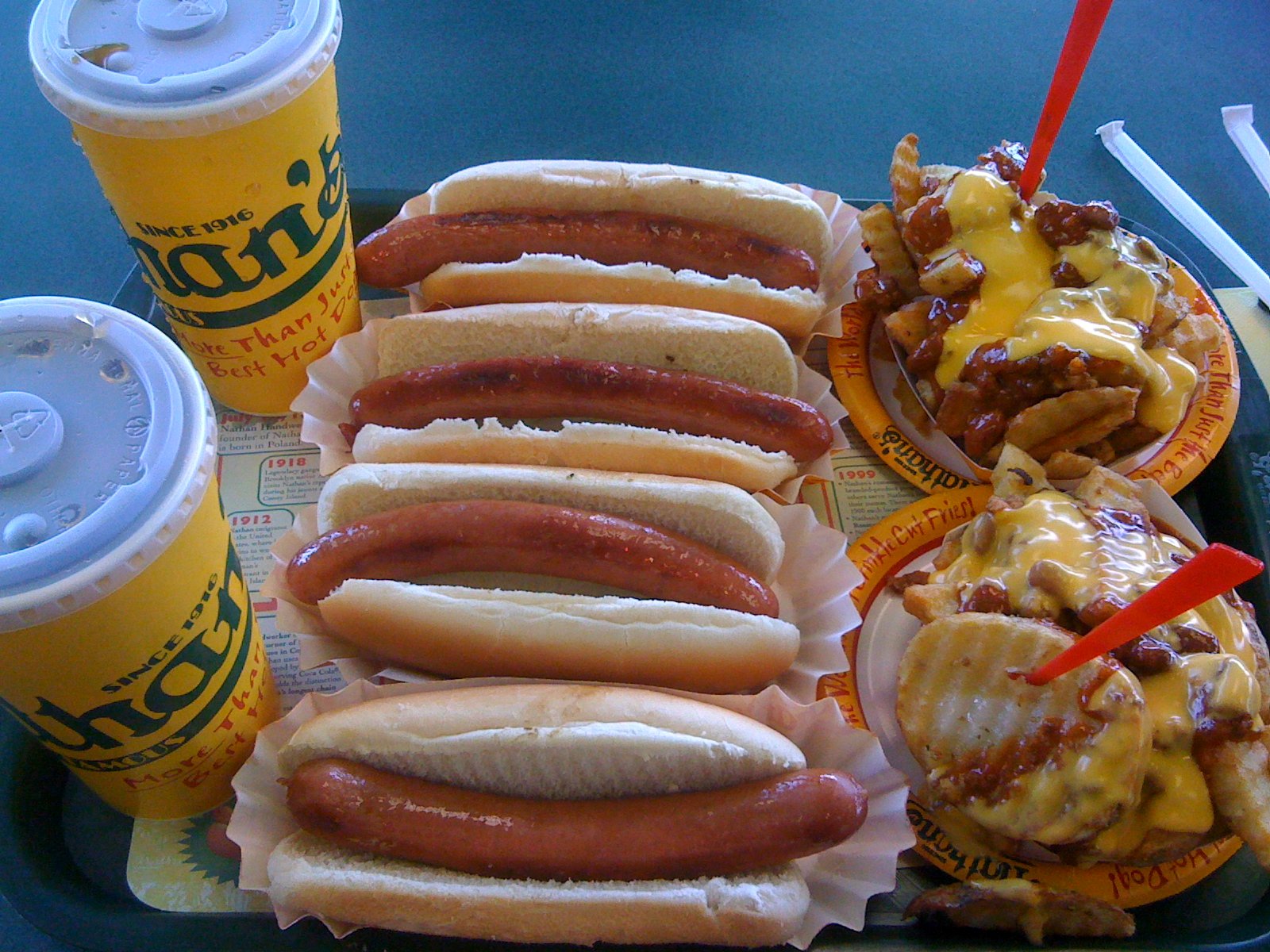 Nathan's のトレーに並ぶホットドッグ4本とチリチーズフライ、ロゴ入りカップ