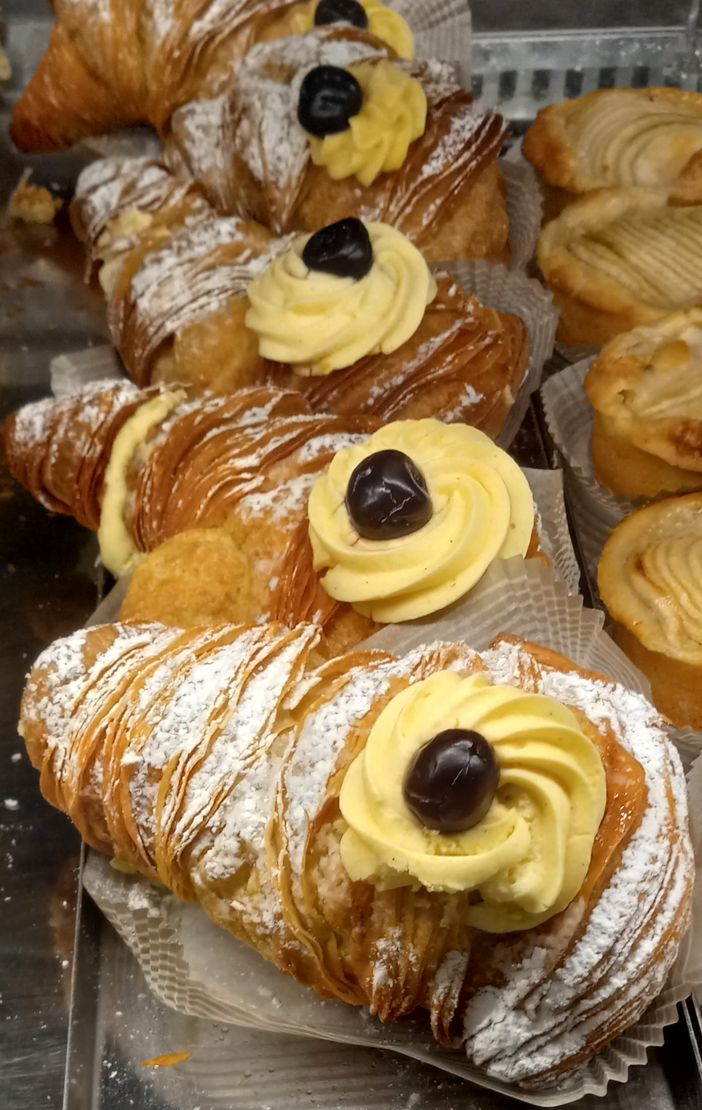 店のケースに並ぶ、カスタードを絞りチェリーをのせたロブスター・テイル（スフォリアテッラ形のパイ）