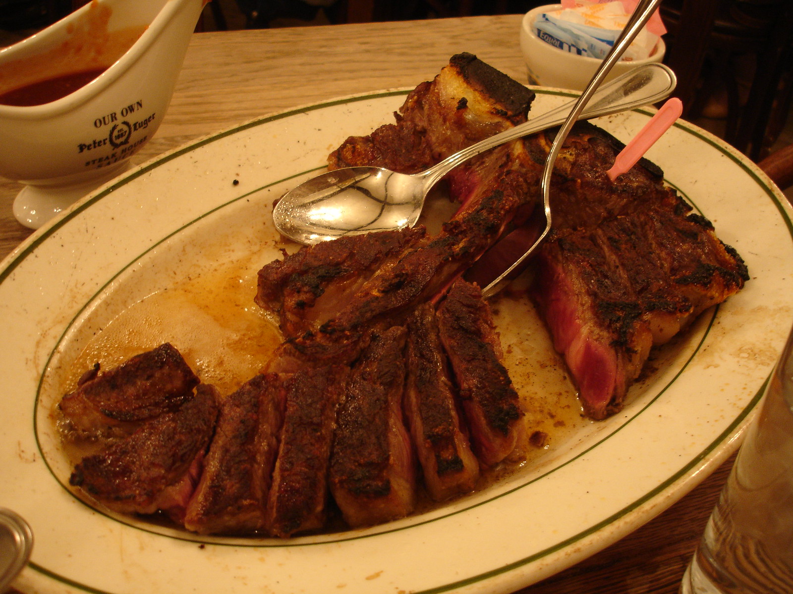 傾けた皿の肉汁の中に並ぶ、焼き色の濃い切り分けたポーターハウス、奥にソースポット
