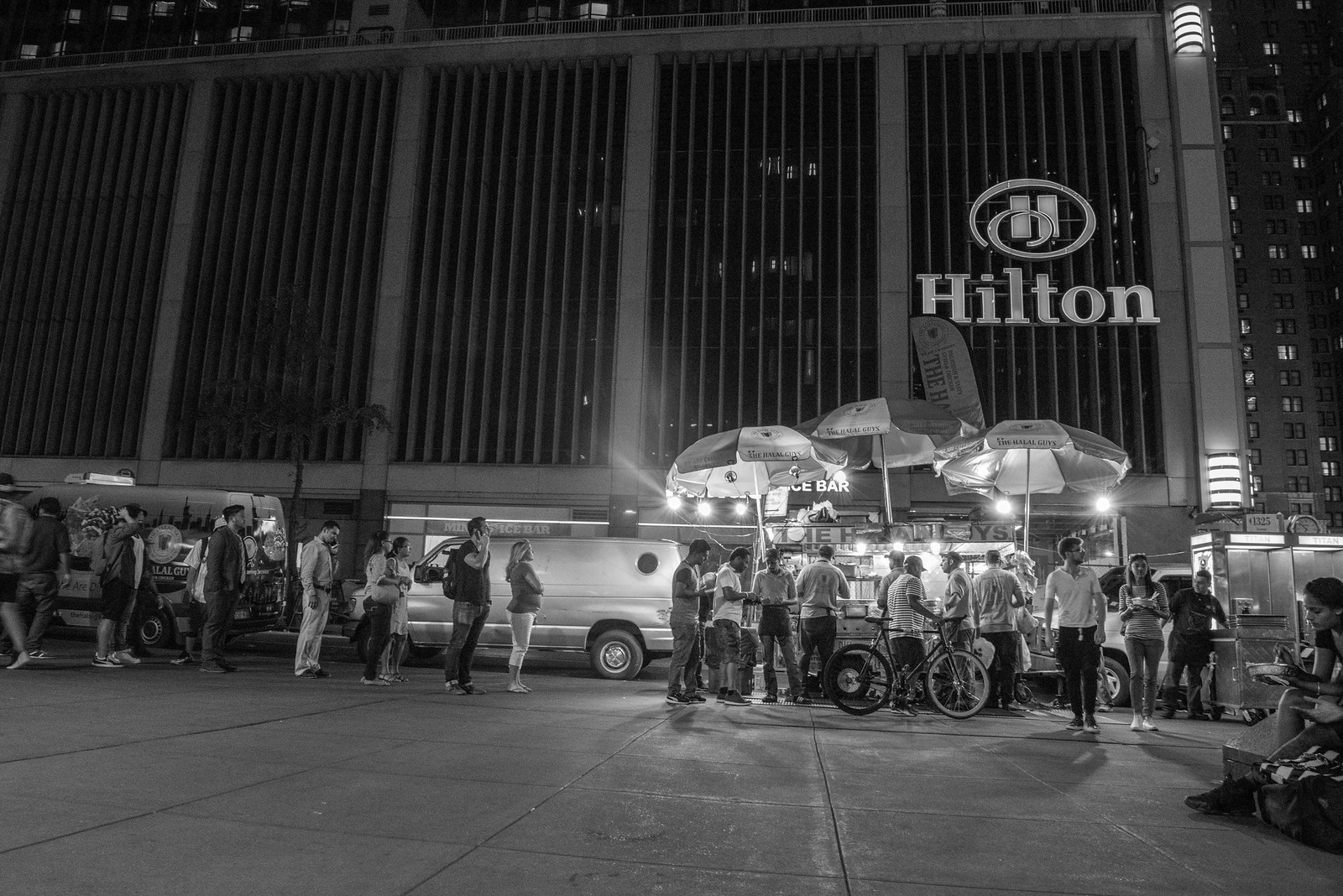 夜のヒルトン前、53丁目と6番街の角に並ぶ The Halal Guys の屋台と行列（モノクロ）