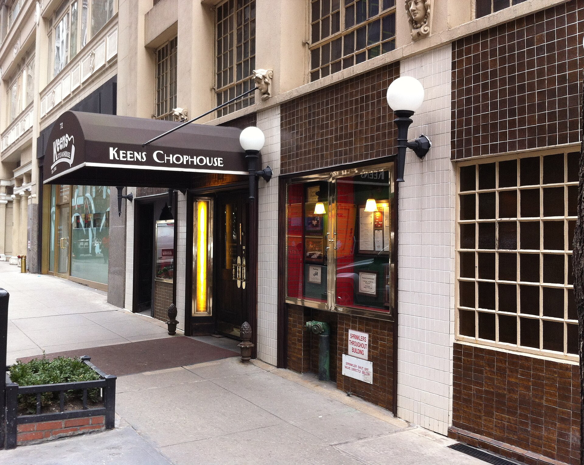 茶色の日よけ「KEENS CHOPHOUSE」と丸いランプが並ぶ店先、昼の歩道