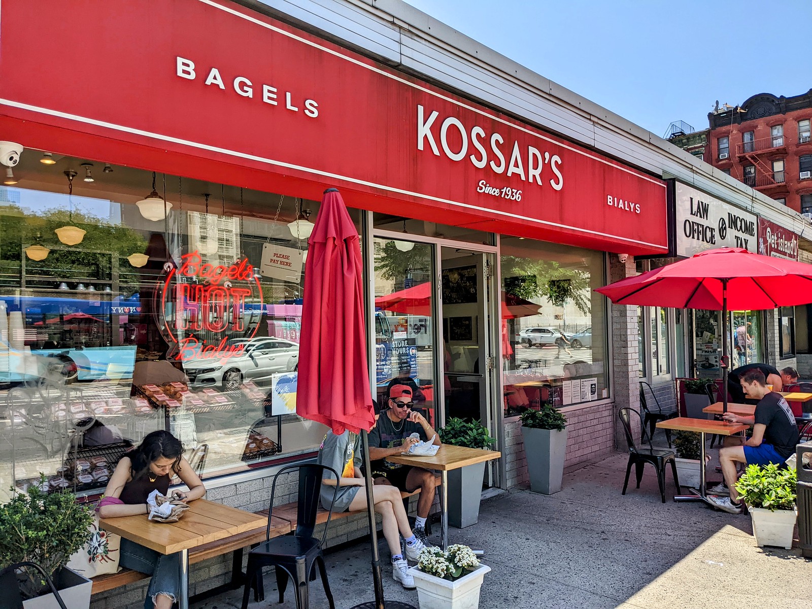 赤い日よけに「KOSSAR'S BAGELS」、歩道に赤いパラソルとテーブル席