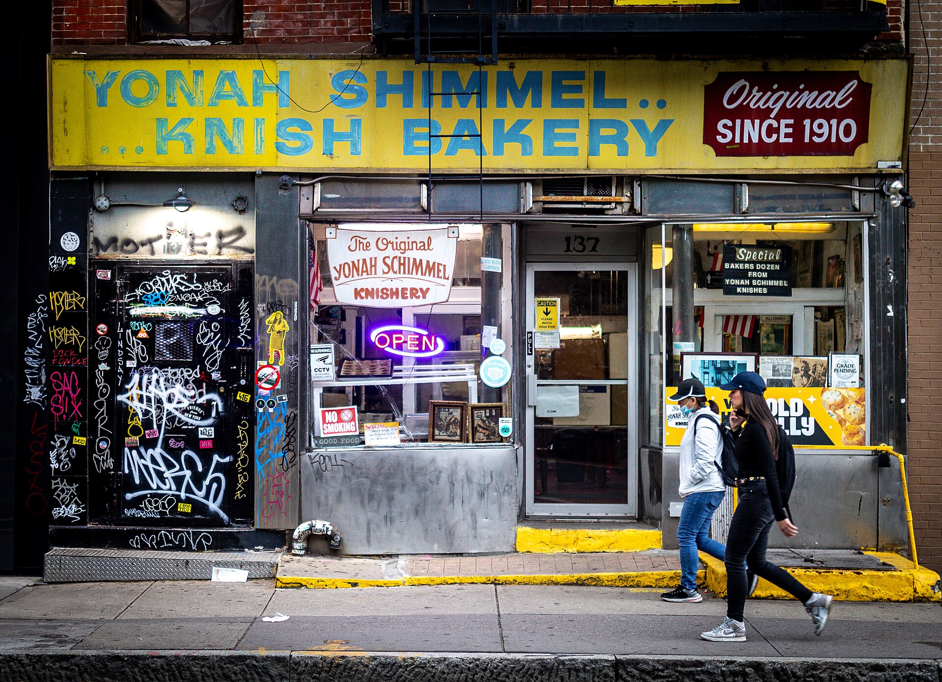 黄色い看板「YONAH SCHIMMEL KNISH BAKERY SINCE 1910」の古い店先と歩道