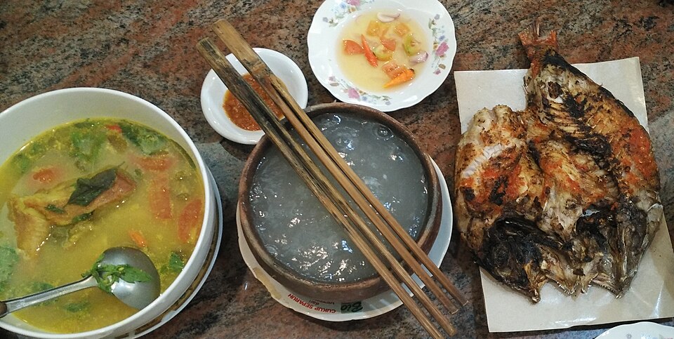 石の卓に並んだ黄色い魚のスープ、木の器に入ったパペダ、焼き魚、サンバル・コロコロ