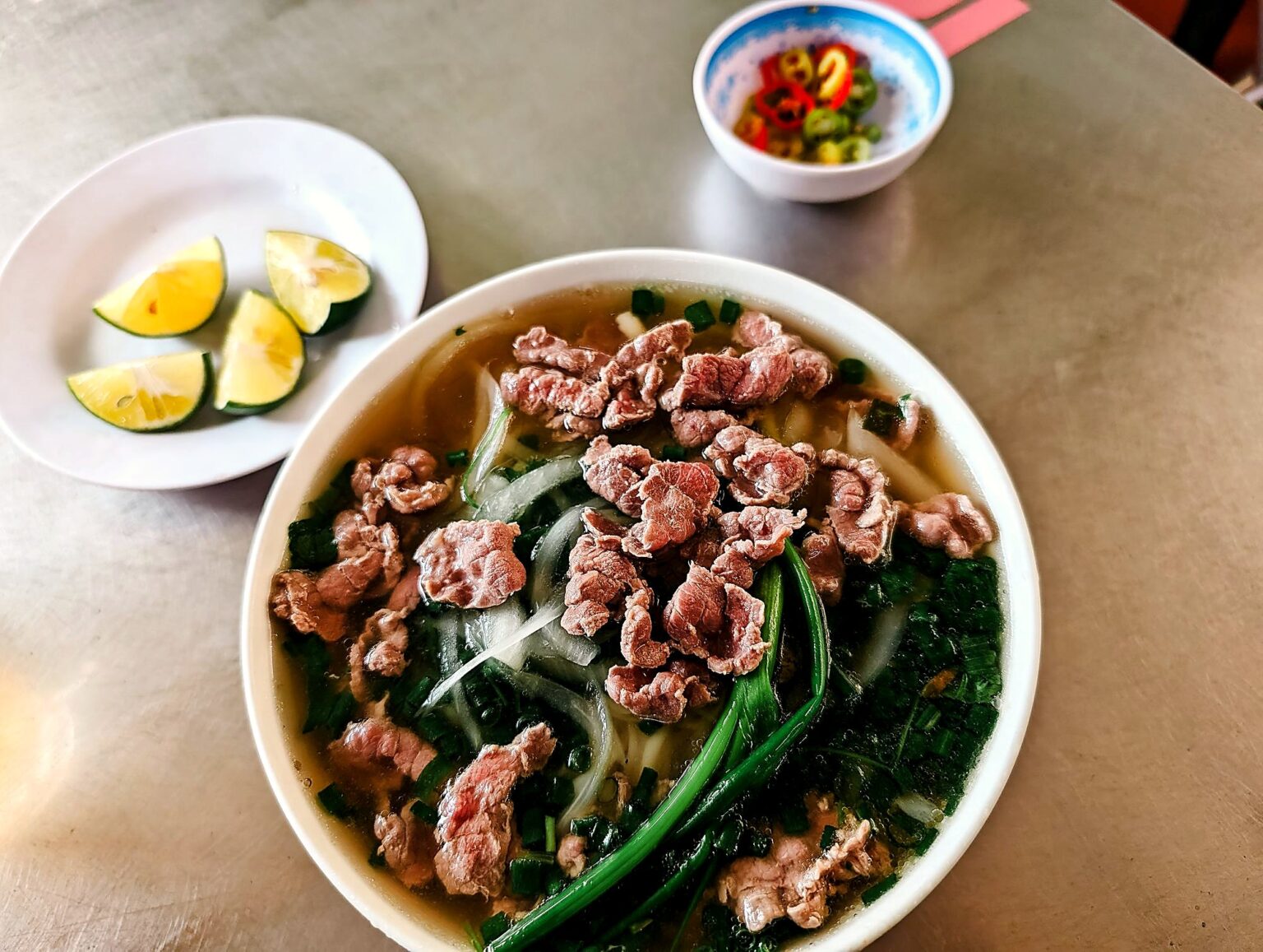 真上から見たフォーの丼。牛肉、青ねぎ、玉ねぎ、ライムと唐辛子の小皿が添えられている