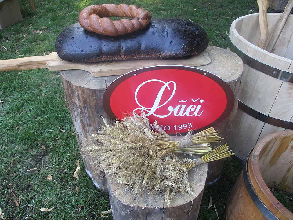 木の台にのせられた、ラーチ製パン所が焼いた大きな黒いライ麦パン。上にソーセージの輪と麦の束が置かれている